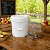 A white plastic scratting bucket with a metal handle sits on a wooden table in an apple orchard setting, with apples in crates
