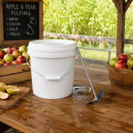 A white plastic scratting bucket with a metal handle sits on a wooden table in an apple orchard setting, with apples in crates