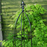 Black metal garden obelisk with decorative top against a green leafy background