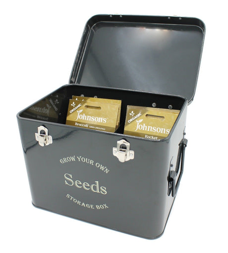 A black metal seed packet storage tin with metal handles, open lid showing compartments with seed packets inside, and text 'GROW YOUR OWN Seeds' on the front.