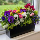 Colorful flower arrangement in a black container on a windowsill with a view of greenery outside.