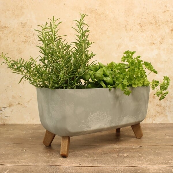 A concrete trough-style plant pot with various herbs planted in it, supported by wooden legs on a wooden surface.