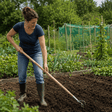 A woman gardening in a lush green vegetable patch, using a wooden-handled garden fork to turn dark soil. She is wearing a navy blue t-shirt, blue jeans, and dark green rubber boots.