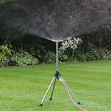 Garden sprinkler on a stand with a hose, spraying water on a grassy area with plants in the background.