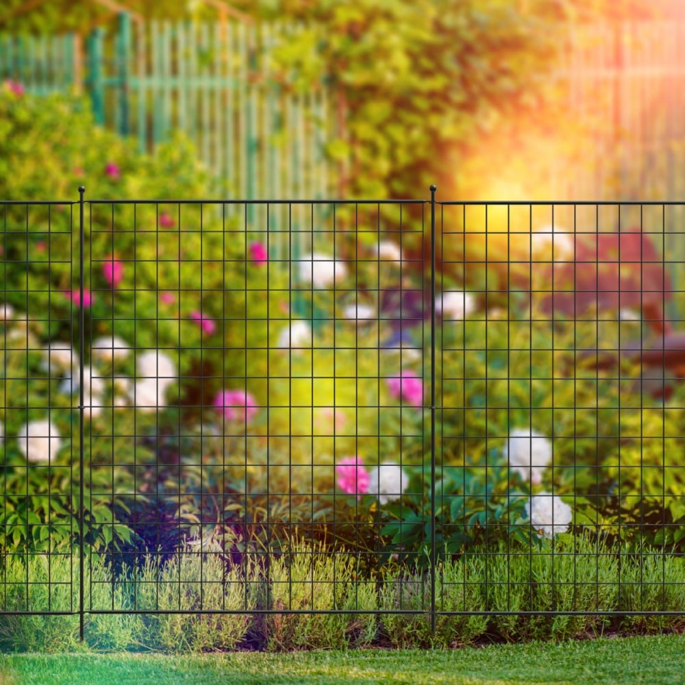 A black metal grid fence panel installed in a garden setting, with a blurred background of colorful plants.