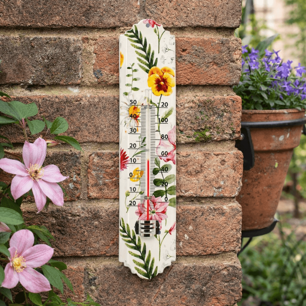 Floral-themed outdoor thermometer on a brick wall with plants in the background
