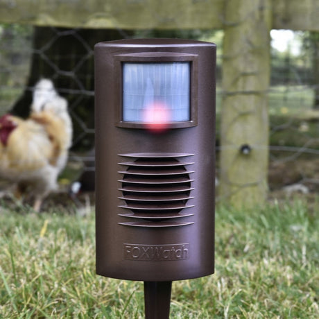 A Foxwatch electronic animal deterrent device, colored brown, with a red indicator light, mounted on a post outdoors.