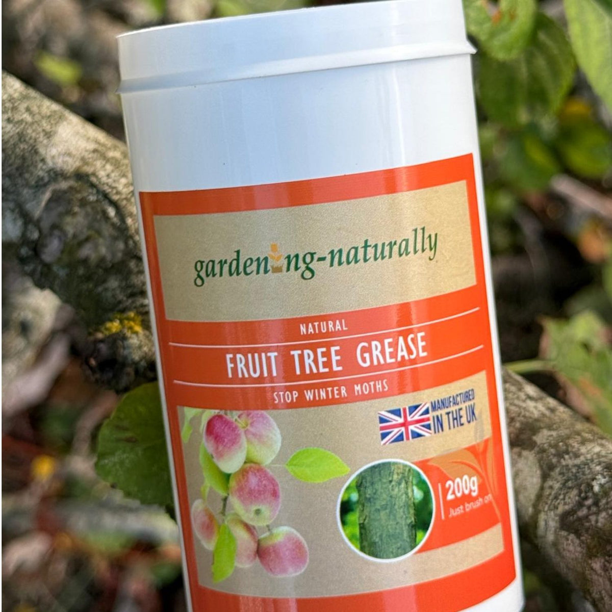 A white plastic tub of gardening naturally Fruit Tree Grease with an orange and beige label, shown outdoors on a tree branch with green leaves in the background.