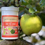 A white plastic tub of gardening-naturally Fruit Tree Grease with a red and beige label, positioned next to a green apple on an apple tree branch in an outdoor garden setting.