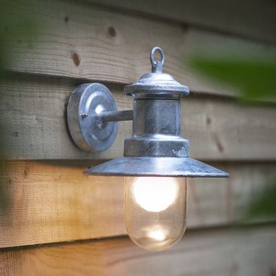 A wall-mounted outdoor light with a galvanised metal finish and a clear glass shade, emitting a warm white light.