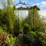 Metal garden archway in a garden setting with plants and a greenhouse in the background.