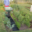 A garden path made of black landscape fabric laid out in a garden bed, with a person in the background holding a wheelbarrow.