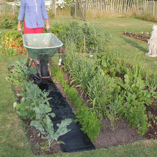 A garden path made of black landscape fabric laid out in a garden bed, with a person in the background holding a wheelbarrow.