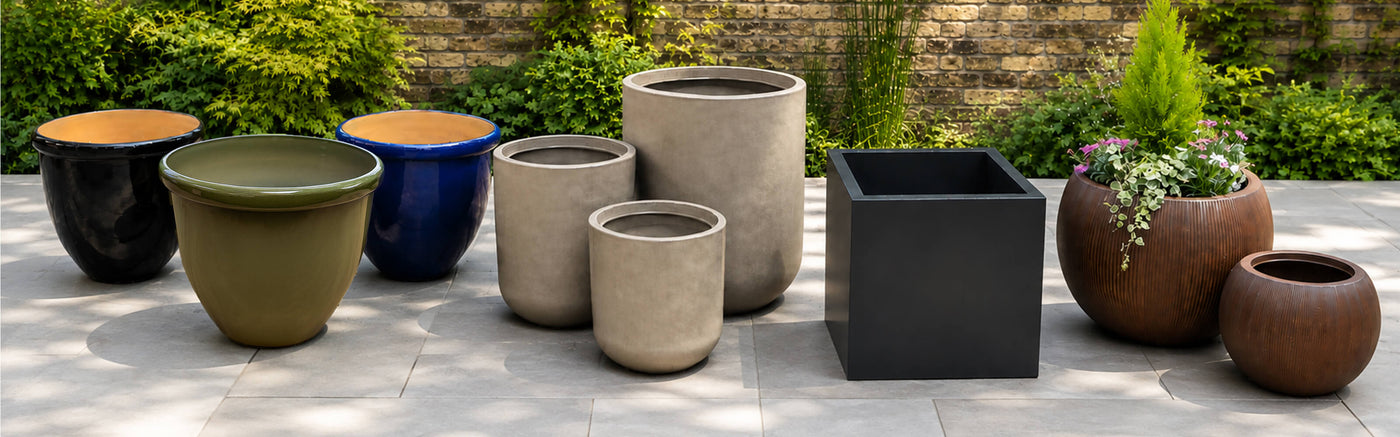 A collection of outdoor plant pots and planters in various shapes, sizes, and colors, including matte black, terracotta brown, olive green, blue, and beige, displayed on a stone patio with greenery and a brick wall background.