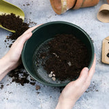 Person sieving soil through a green mesh container on a concrete surface.