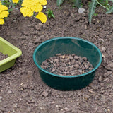 Green garden sieve filled with soil on a garden bed with yellow flowers.