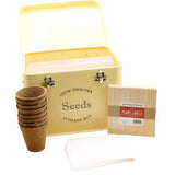 A cream-colored metal seed storage box with wooden plant labels, brown plant pots, and seed envelopes.