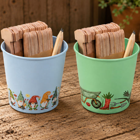 Two decorative gardening tins, one light blue with gnome illustrations and one mint green with gardening tools, each filled with wooden plant labels and a pencil, set on a rustic wooden table.