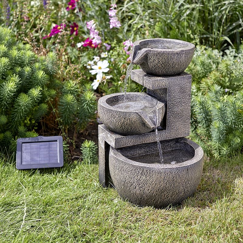 A stone effect water feature with a solar panel positioned on grass surrounded by flowers, showing water flowing from the top bowl into the bowl below.