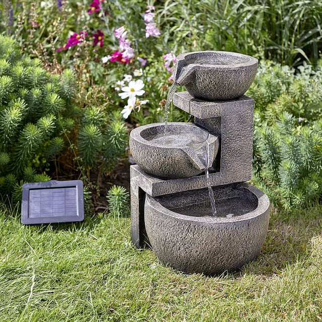 A stone effect water feature with a solar panel positioned on grass surrounded by flowers, showing water flowing from the top bowl into the bowl below.
