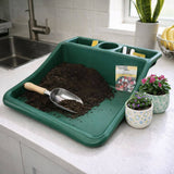 Green gardening tray with soil, a scoop, and small plants on a kitchen counter.
