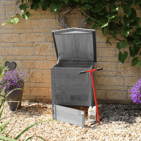 Gray beehive composter with a red tool on a gravel surface against a brick wall.