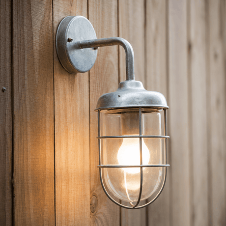 A wall-mounted garden light with a galvanised steel cage and a lit bulb, attached to a wooden surface.