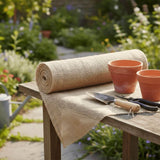 A roll of natural burlap garden matting draped over a wooden table in a sunny garden, with terracotta pots, a trowel, and pruning shears placed on top.