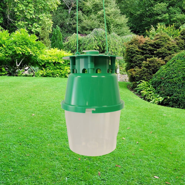 A green and translucent plastic hanging trap, likely for mosquitoes or insects, suspended by green cords in a lush green backyard setting.