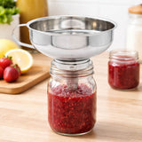 Metal funnel being used to transfer red jam into a glass jar on a wooden surface with fruits in the background.