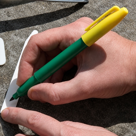 A person's hands holding a garden label marker pen with a green body and yellow cap, writing on a white label.