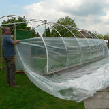 Person installing clear plastic sheeting over a greenhouse structure on a grassy area.