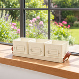 Three cream-colored herb tins, arranged in a row on a wooden windowsill with a blurred garden background.