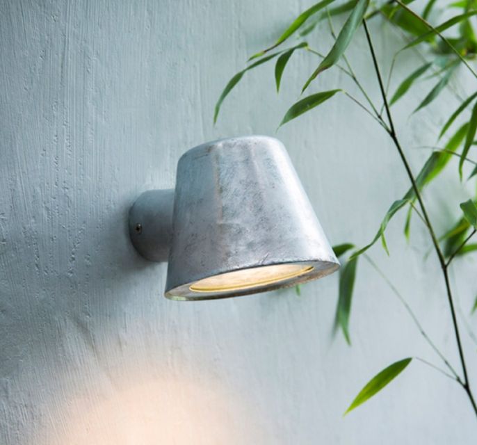 A wall-mounted outdoor light fixture with a galvanised steel finish, emitting a warm white glow, positioned beside a green potted plant.