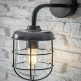 A dark grey garden wall light with a nautical cage design, mounted on a wall with a visible brick pattern.
