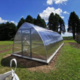 A polytunnel greenhouse with a metallic structure and clear polycarbonate panels, situated outdoors with a mountainous landscape in the background.
