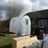 Small greenhouse in a garden with a wooden planter and a house in the background.