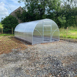 A polytunnel greenhouse with a metallic frame and clear polycarbonate covering, situated outdoors with a fence in the background.
