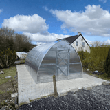 Metal greenhouse on a concrete base with a house and trees in the background