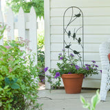 A black metal pot trellis with perching bird designs is displayed behind a potted plant on a patio, with green plants in the background.