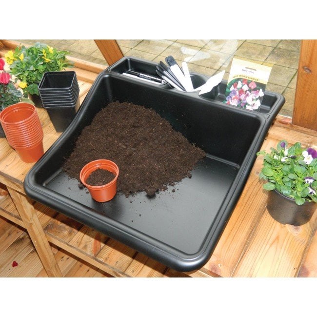 A black potting tray filled with soil, placed on a wooden table with various gardening tools and plants around it.