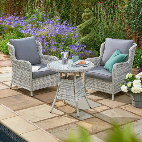 An outdoor garden bistro set with two high-back chairs and a glass-topped table, placed on a patio with flowers in the background.