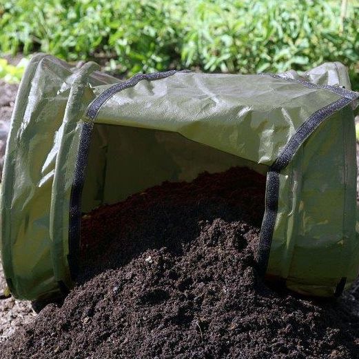 A green rolling composter made of fabric, placed on the ground with soil spilling out of it, indicating it is full of compost.