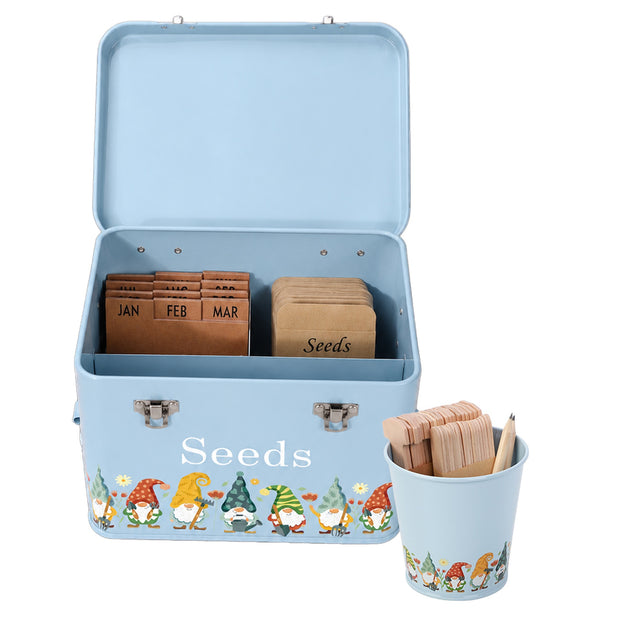 A light blue metal seed storage tin with an open lid, decorated with a row of colorful garden gnomes and the word 'Seeds' on the front. Inside, wooden seed tabs by months and a wooden 'Seeds' tab are organized.