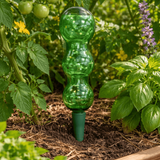 Green plastic water dispenser in a garden setting with plants and flowers.