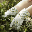 A pair of white gardening rigger gloves with green leaf patterns, worn by a person amidst green foliage.