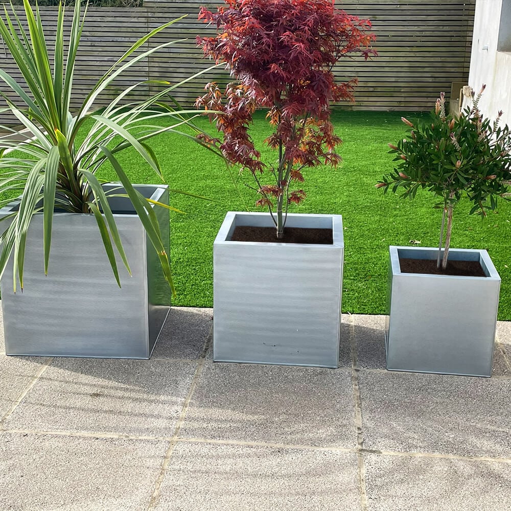 Three silver cube-shaped zinc planters in different sizes, with the smallest in front, medium in the middle, and largest in the back, placed on a patio with plants inside them.