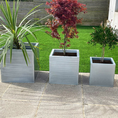 Three silver cube-shaped zinc planters in different sizes, with the smallest in front, medium in the middle, and largest in the back, placed on a patio with plants inside them.