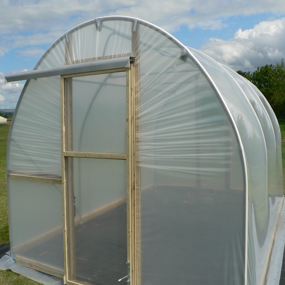 Small greenhouse with transparent plastic covering on a grassy area with trees in the background