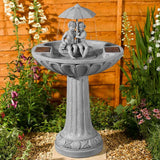 A gray stone-style garden bird bath fountain with a pedestal base, a shallow round basin, a small sculpted figure sitting under a rain umbrella, and solar panel covers on the sides, set outdoors in front of a wooden fence with plants and flowers.